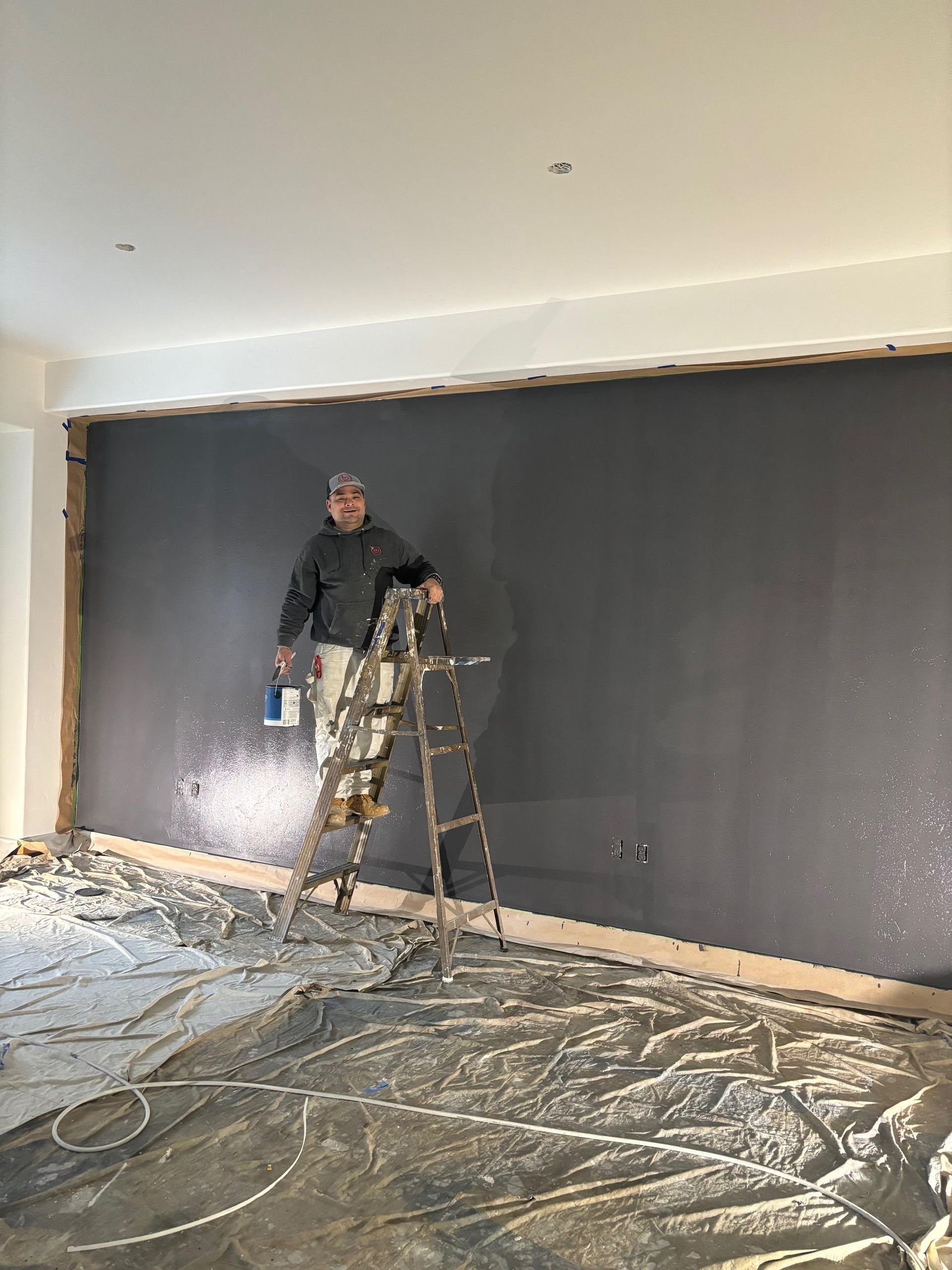 A painter on a step ladder finishing a charcoal accent wall, floor covered with drop cloths