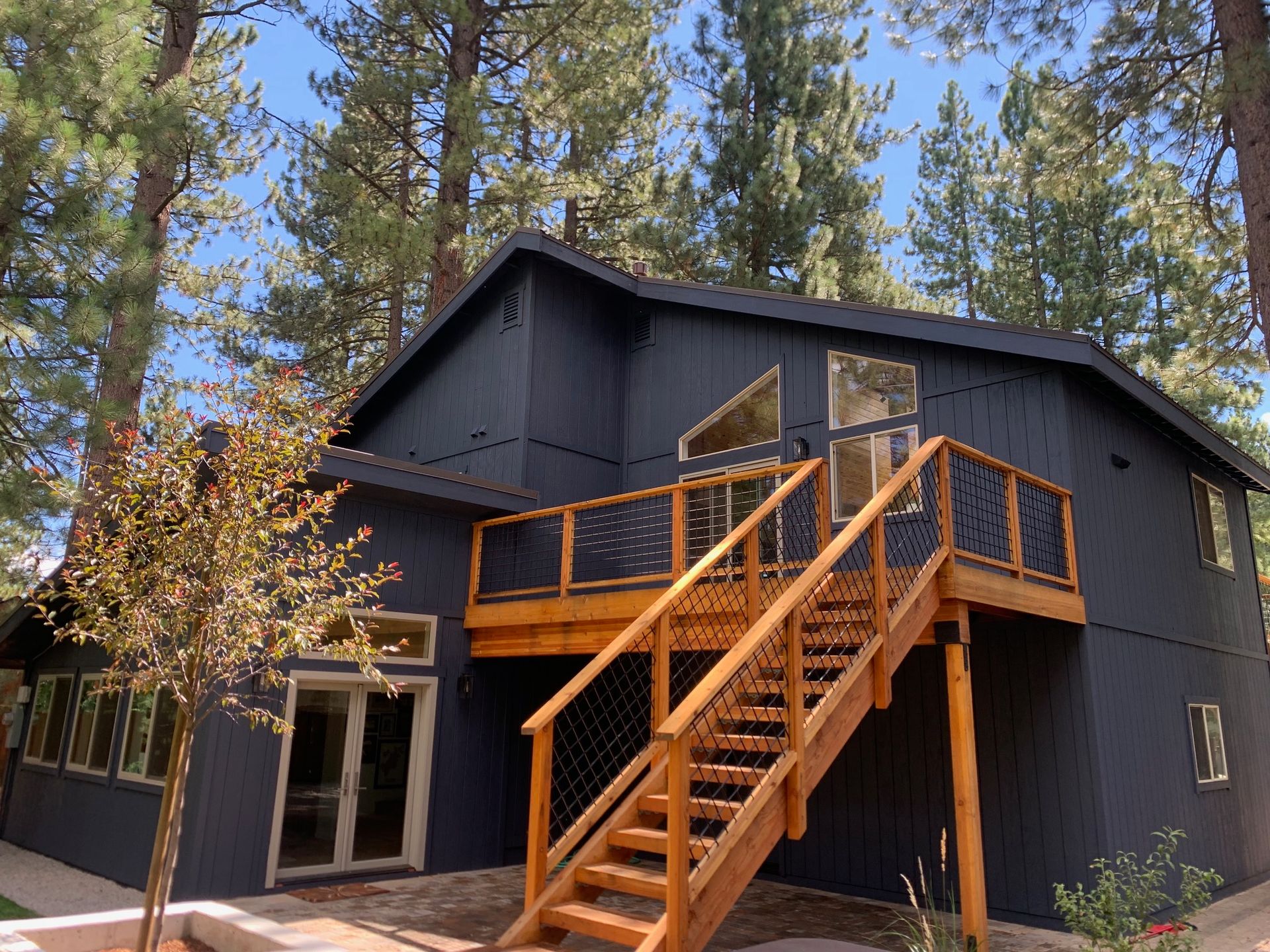 Exterior repaint in deep charcoal with a new cedar deck and stair rail, surrounded by pines